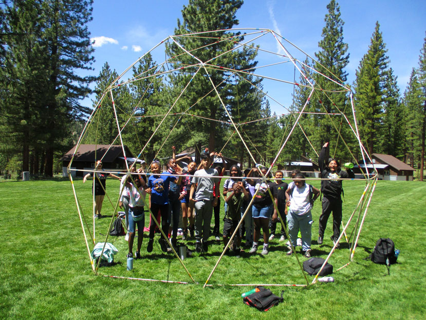 A group of kids (late elementary school aged) stand outside in the forest and are inside a hexagonal dome held together by thin pieces of metal arranged in large triangles that are large enough for the kids to bend over and walk through