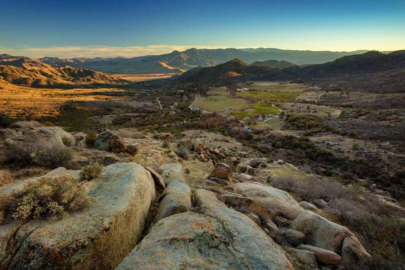 looking out and down across a rocky hillside with clusters of shrubs and small trees leading to a few dwellings at the base and mountains in the background