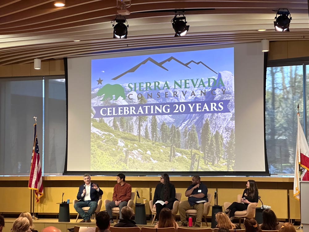 A group of five people are sitting on stage with a large pull down screen behind them with the Sierra Nevada Conservancy logo projected on it