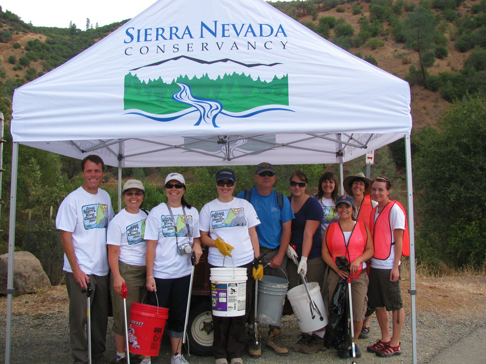 ten people holding buckets and trash picker sticks stand under tent with the Sierra Nevada Conservancy logo on it and a brown hillside with green bushes in the background