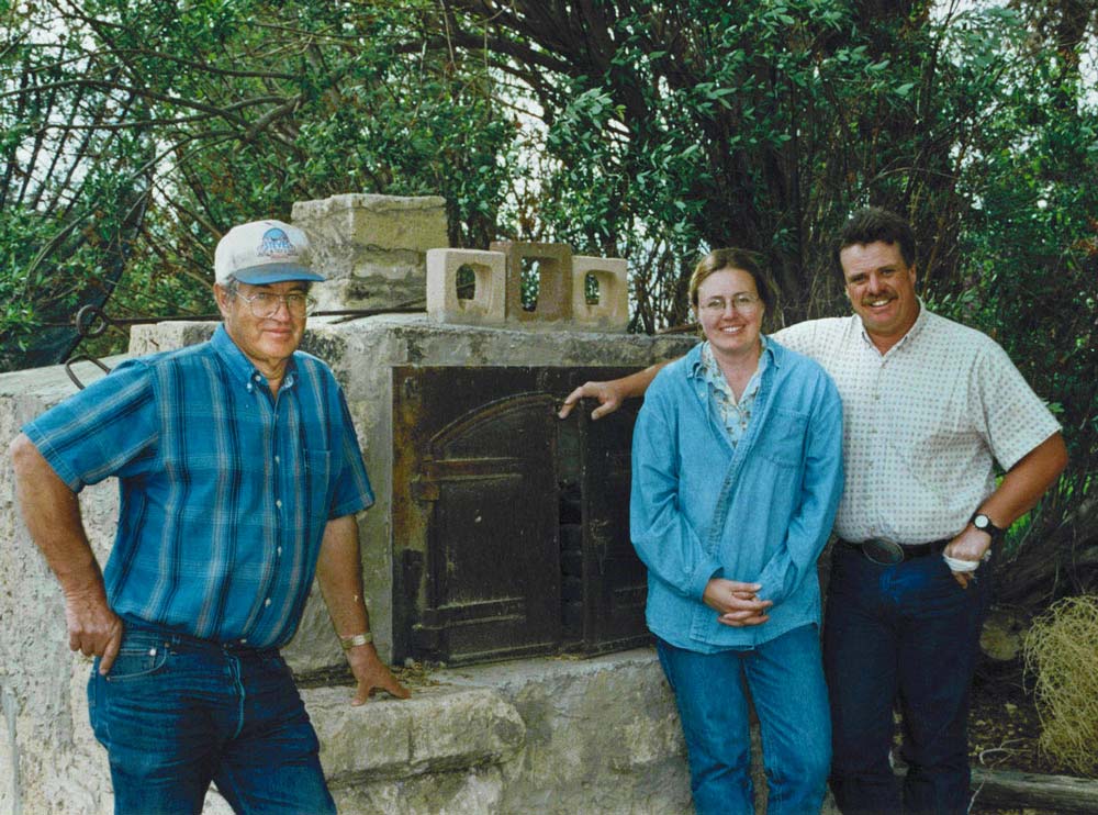 A white man in in sixties, a white woman in her thirties, and white man with a mustache in his thirties stand outside in front of an old rock and metal oven