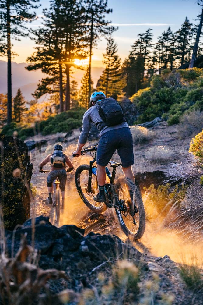 two people mountain biking on a dirt trail with the rocky mountainside to their right with lots of green trees as the evening sun starts to set