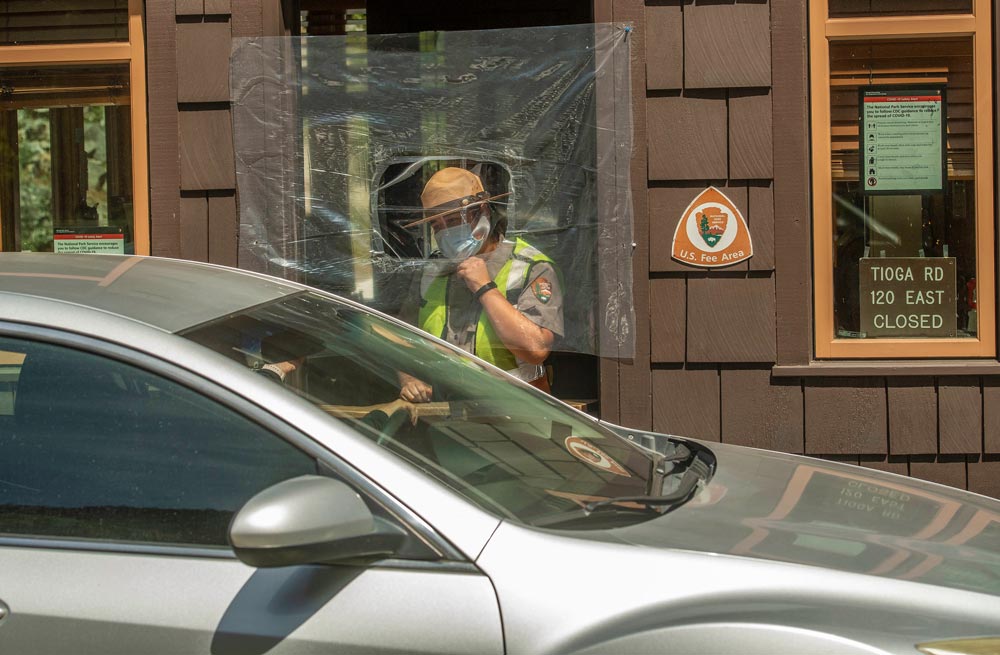 A car is stopped at a booth talking to an attendant, who is standing in the doorway behind a plastic shield wearing a ranger hat and face mask.