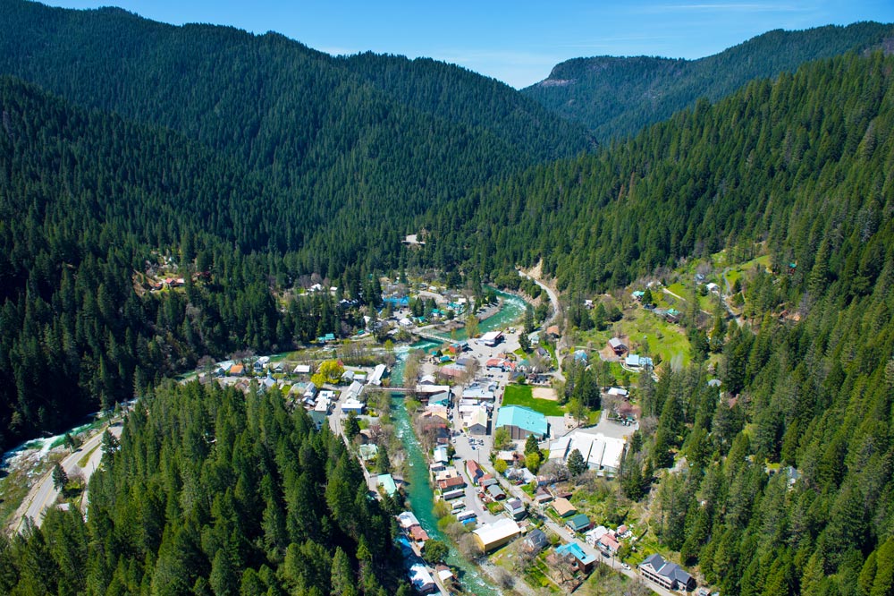 aerial looking down between mountains covered in green trees to a town with several buildings along a river that winds through
