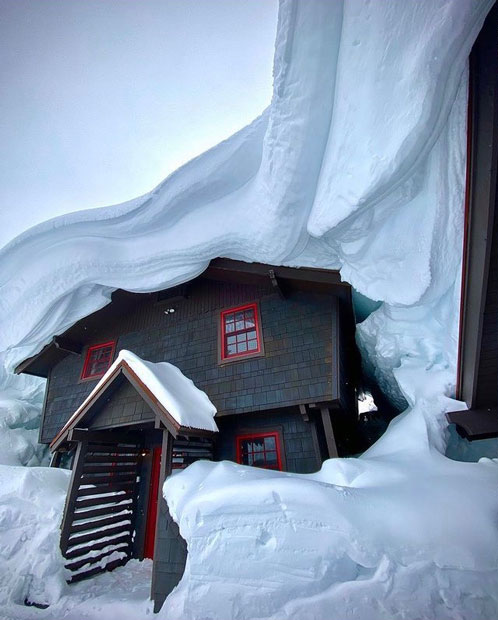 a home engulfed in snow: several feet of snow cover the roof and several feet of snow surround it on all other sides